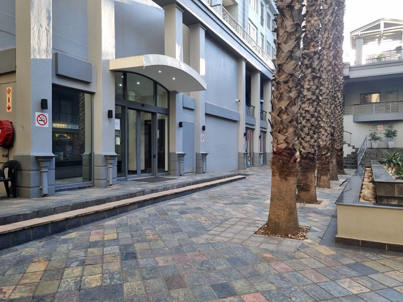 Exterior view of the apartment building entrance featuring paved courtyard, modern glass sliding doors, and decorative palm trees.