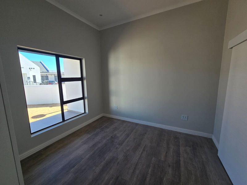 A bright, empty bedroom featuring modern laminate flooring, neutral walls, and a large aluminium-framed window providing natural light.