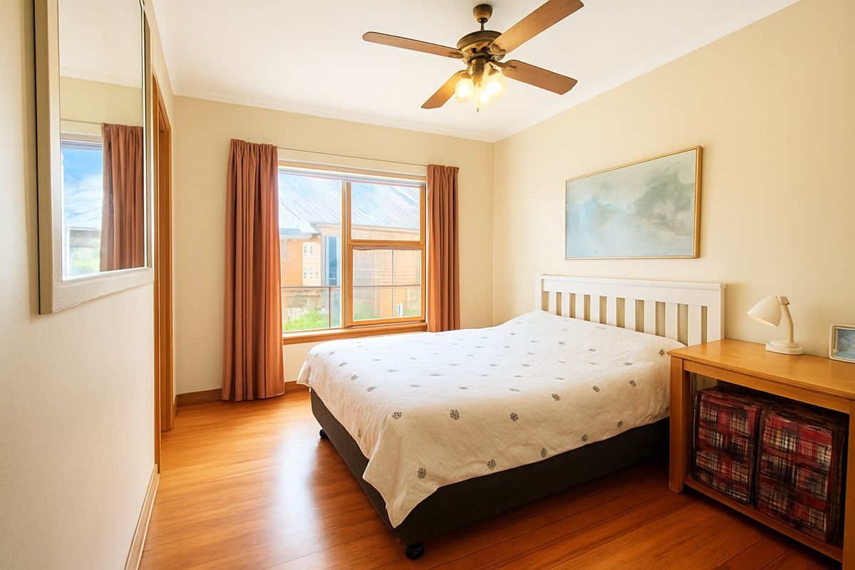 A well-maintained bedroom featuring warm timber flooring, a ceiling fan, and large windows that provide natural light.