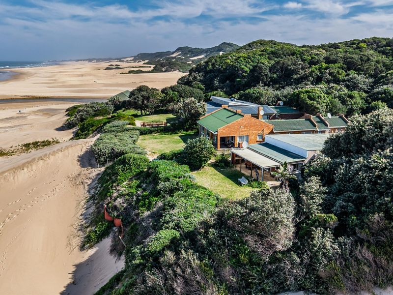 Aerial view of a coastal property featuring a brick house with a metal roof, situated on a dune overlooking the beach and ocean.