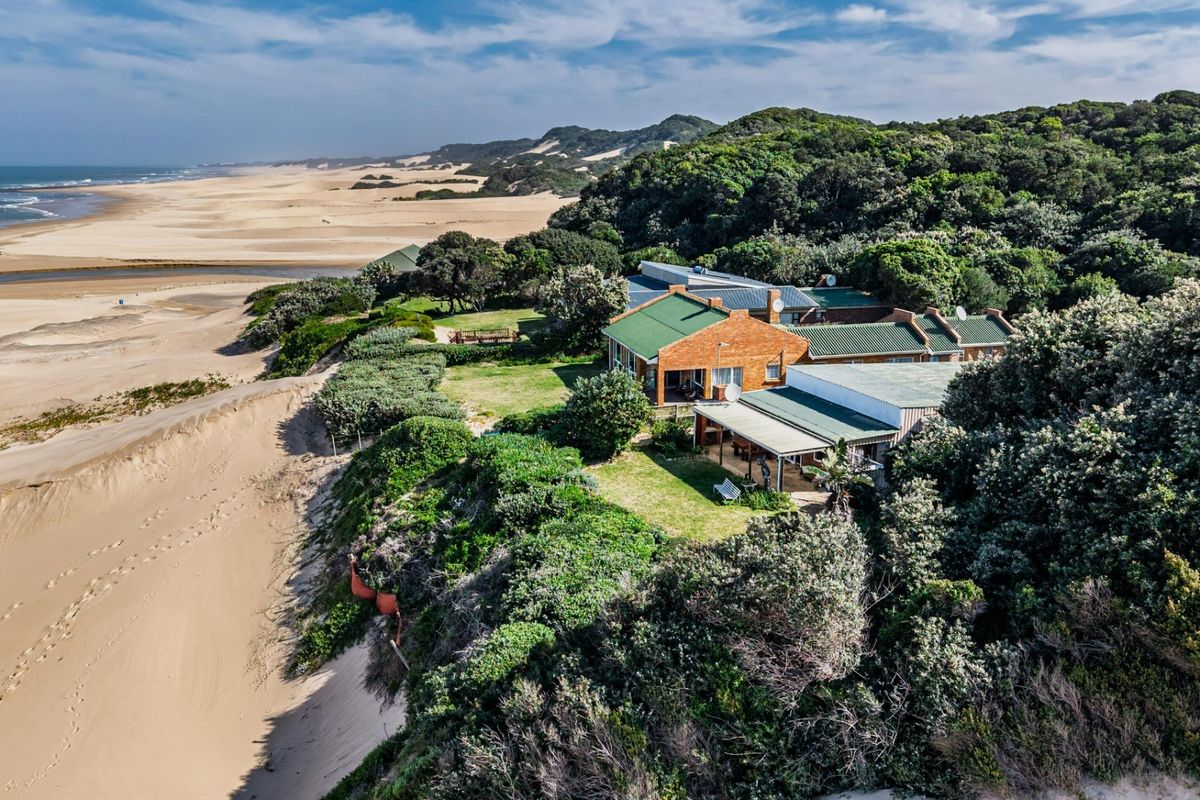 Aerial view of a coastal property featuring a brick house with a metal roof, situated on a dune overlooking the beach and ocean.