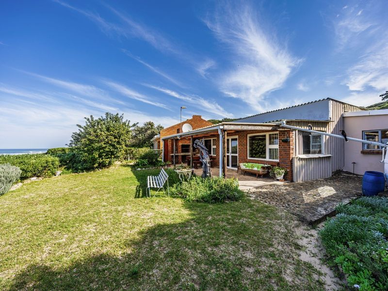 Rear view of the property featuring a brick facade, covered patio area with built-in braai, and a water tank, set within a coastal garden.