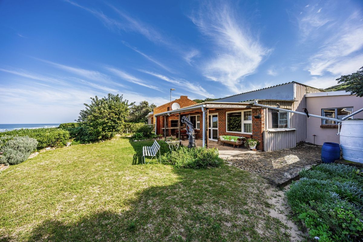 Rear view of the property featuring a brick facade, covered patio area with built-in braai, and a water tank, set within a coastal garden.