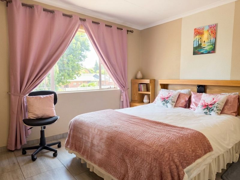 A well-lit bedroom featuring tiled flooring, a built-in wooden headboard with integrated shelving, and a large window with curtain fixtures.