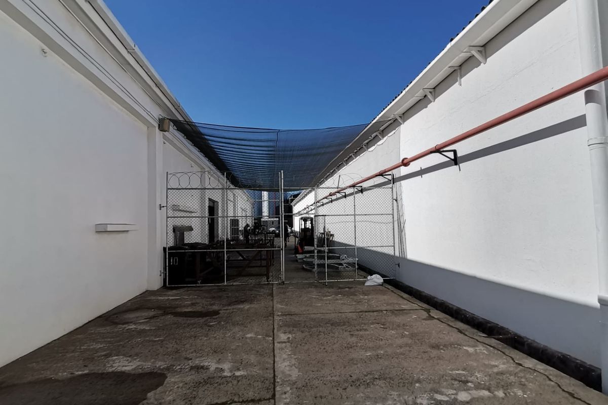 An industrial outdoor corridor between two warehouse buildings, featuring concrete paving, perimeter fencing, and overhead shade netting.
