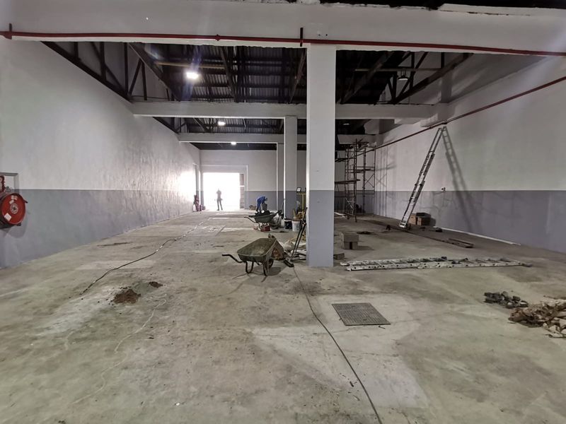 Spacious industrial warehouse interior featuring high ceilings, exposed structural beams, fire suppression piping, and concrete flooring, currently undergoing renovation.