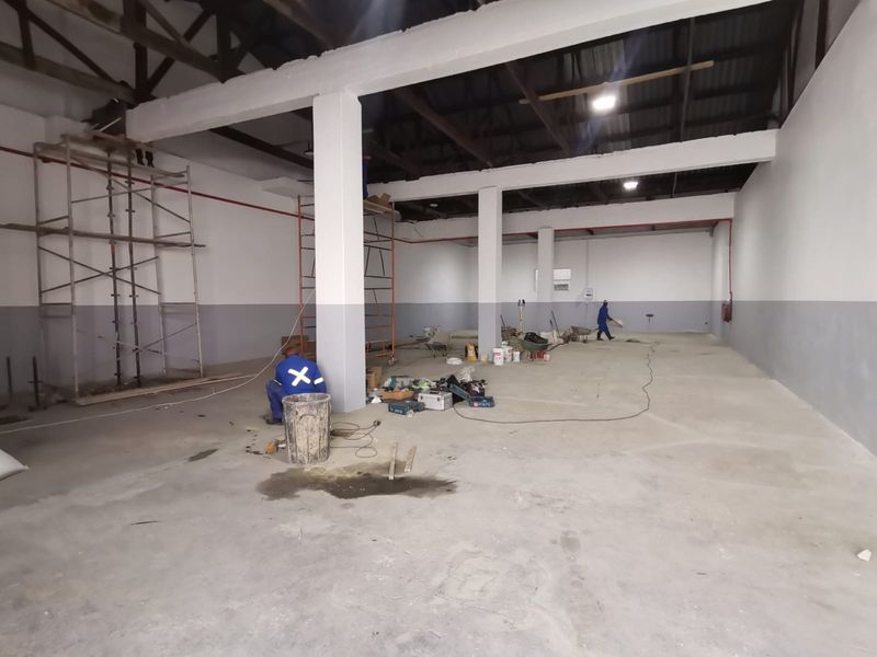 Spacious industrial warehouse interior featuring high ceilings, exposed steel roof trusses, concrete flooring, and structural support columns, currently undergoing maintenance and painting.
