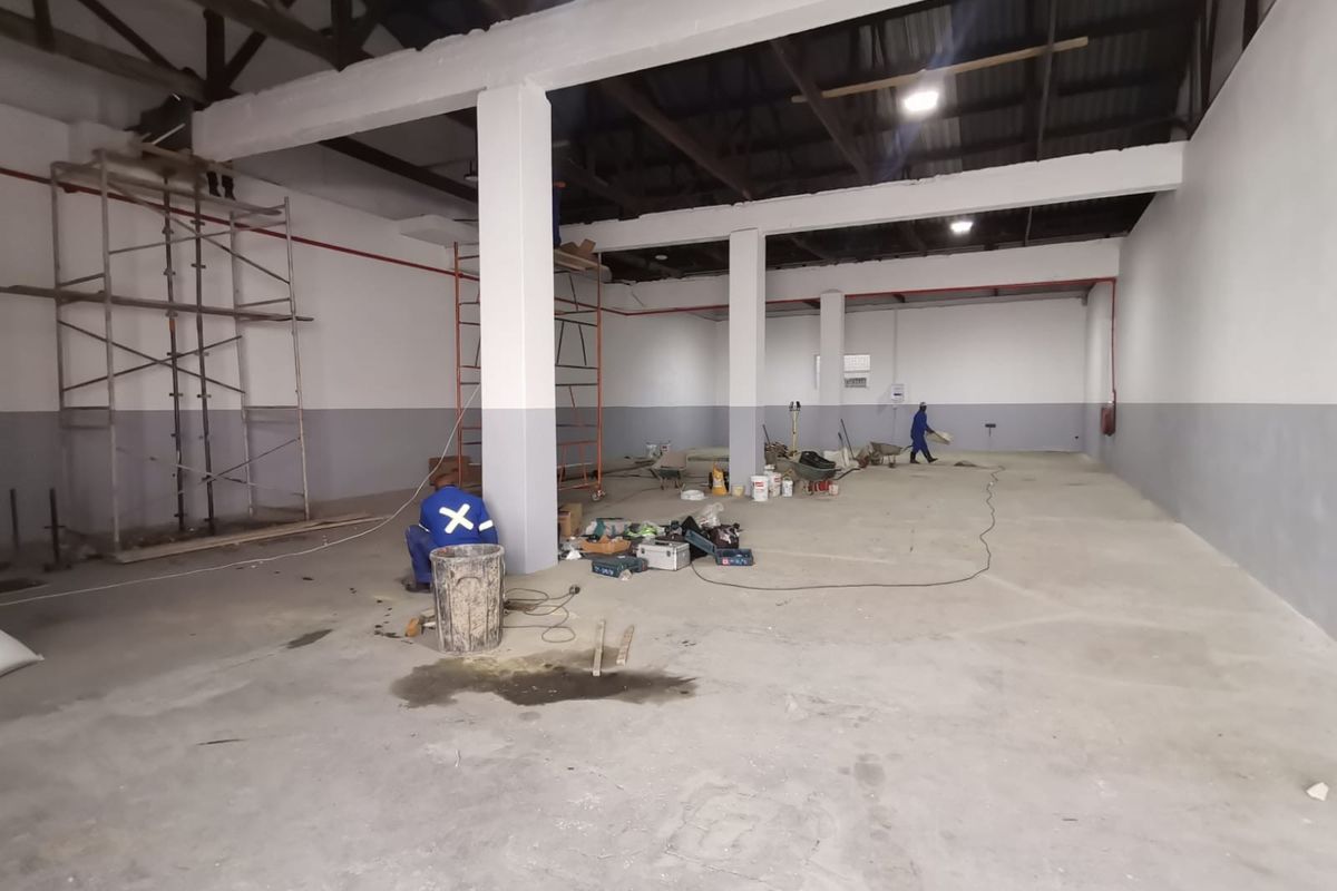 Spacious industrial warehouse interior featuring high ceilings, exposed steel roof trusses, concrete flooring, and structural support columns, currently undergoing maintenance and painting.