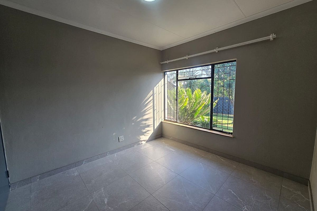 A bright, empty bedroom featuring neutral grey walls, modern tiled flooring, and a large window with security bars overlooking the garden.