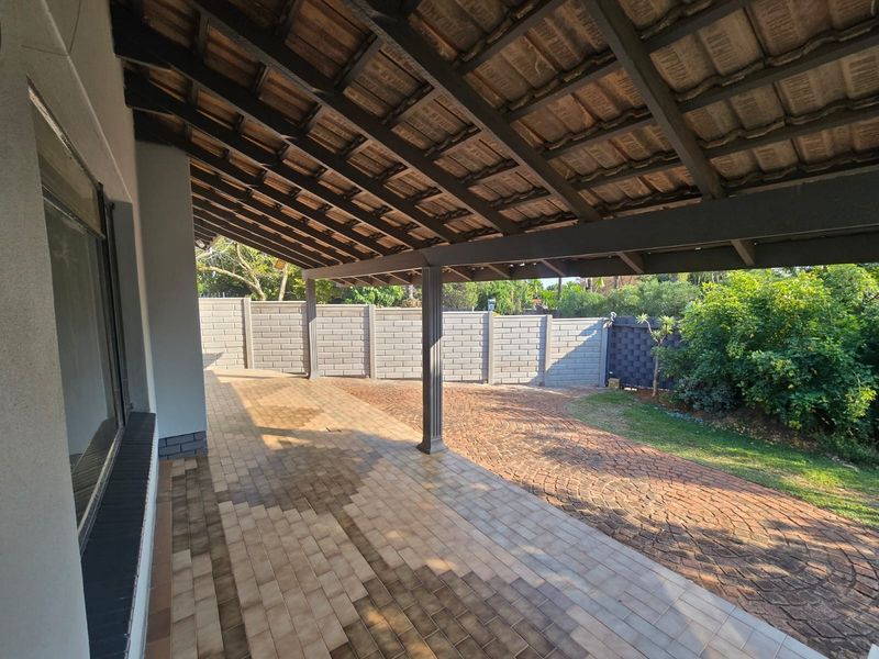 A spacious covered patio featuring tiled flooring and exposed timber rafters, overlooking a paved backyard area enclosed by a brick boundary wall.