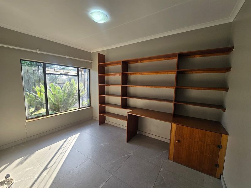 A bright, empty office space featuring built-in wooden shelving and desk units, large windows with security bars, and modern tiled flooring.