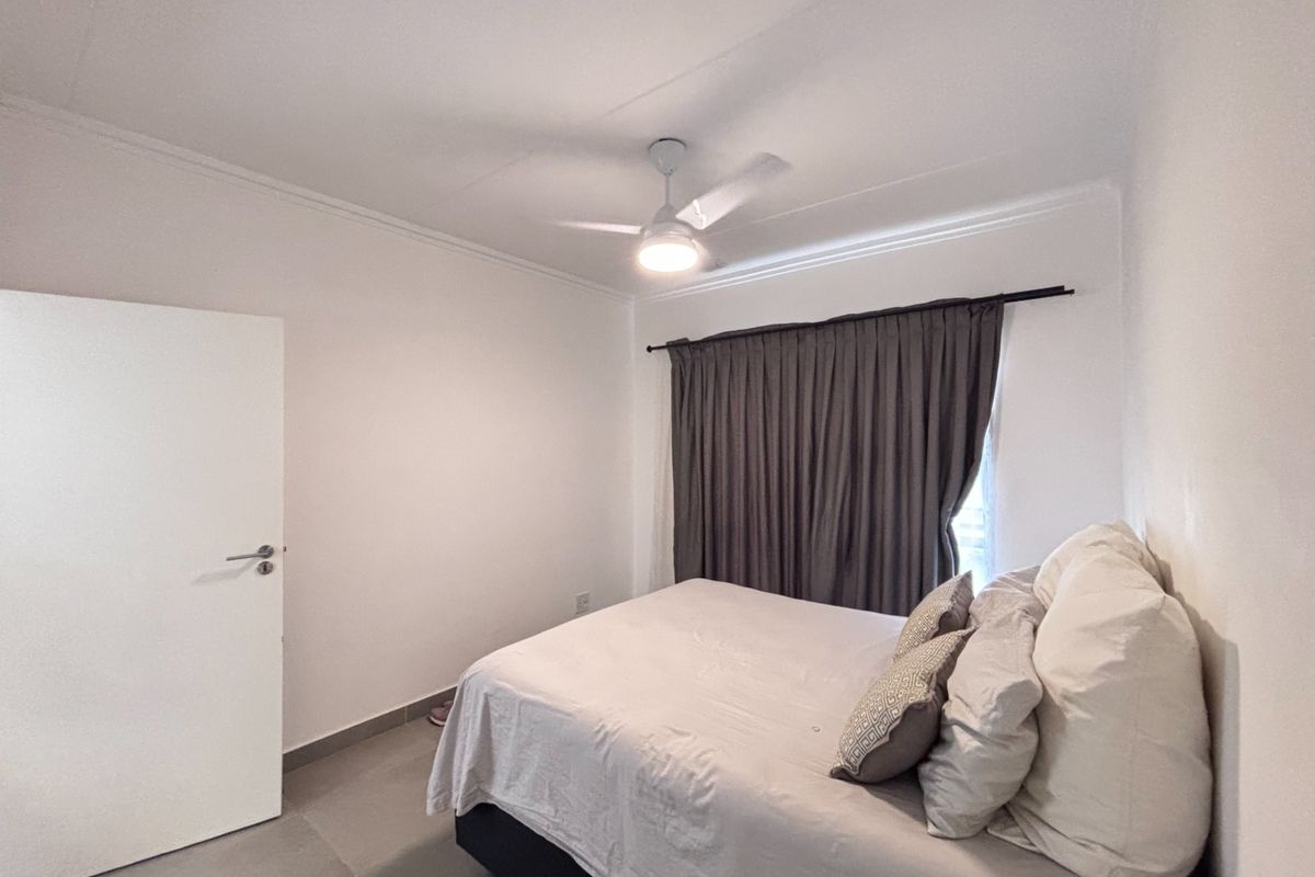 A clean and well-maintained bedroom featuring neutral tiled flooring, a ceiling fan with integrated lighting, and a window fitted with dark curtains.