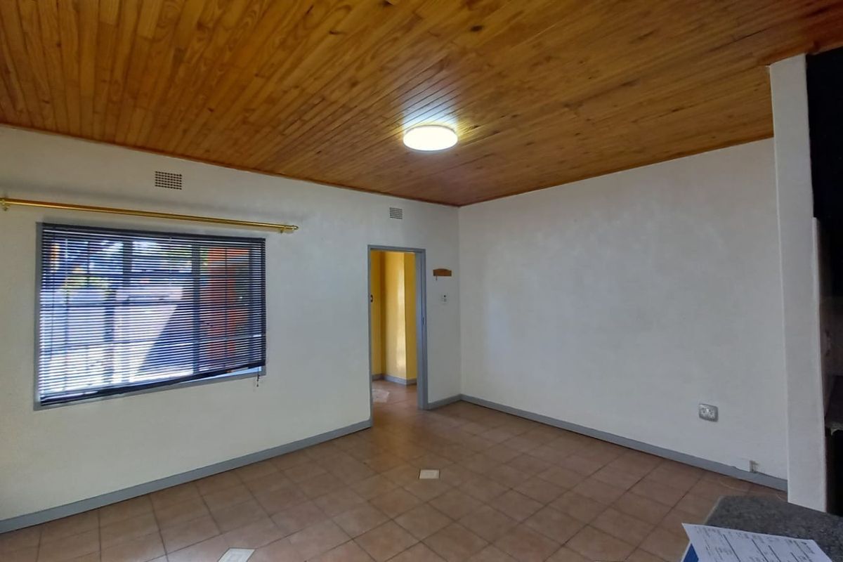 An empty living area featuring a timber-clad ceiling, tiled flooring, and a window with fitted blinds, leading to an adjacent room.