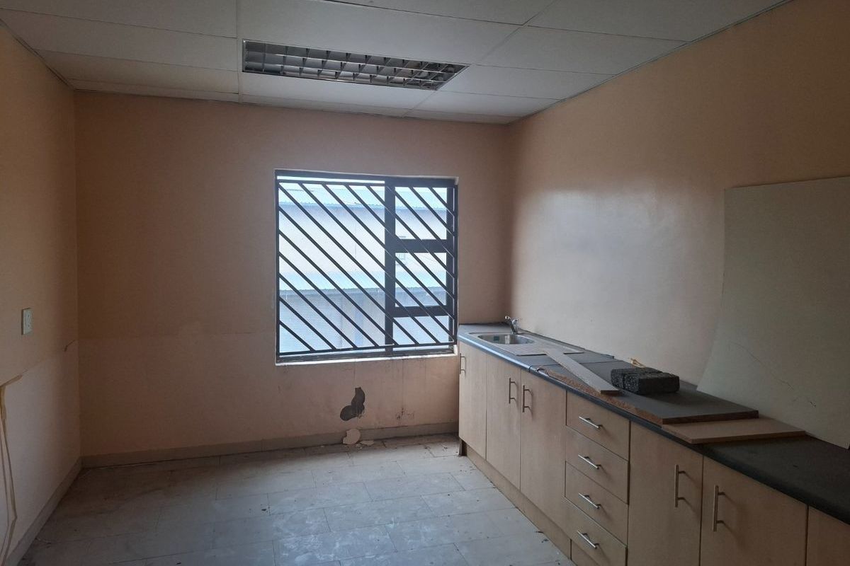 A small commercial kitchenette featuring built-in cabinetry, a sink, and security bars on the window, requiring cosmetic updates.