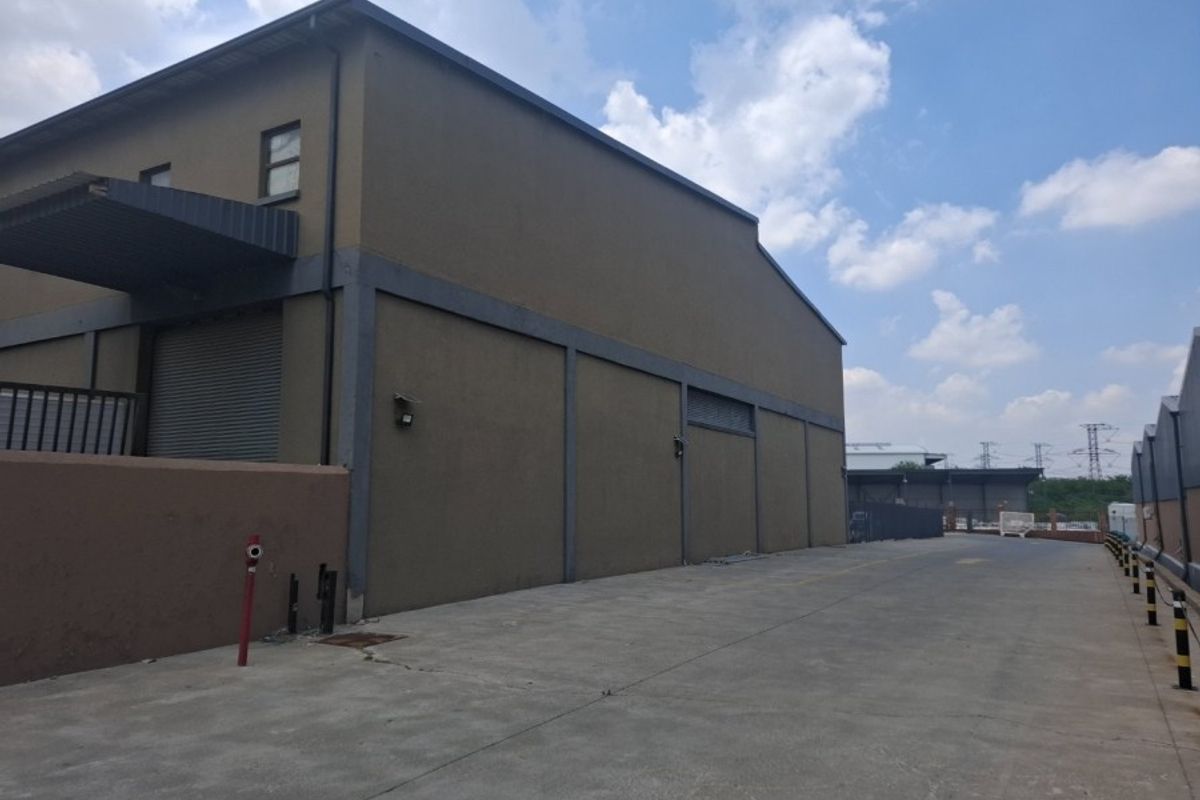 Rear exterior view of a commercial warehouse building featuring a concrete driveway, roller shutter access, and perimeter security infrastructure.
