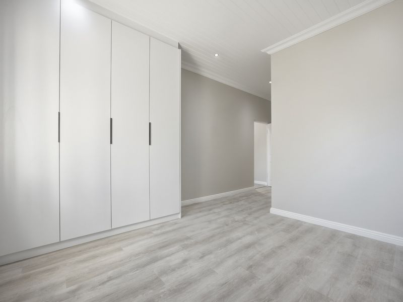 A bright, modern bedroom featuring neutral walls, light laminate flooring, built-in cupboards, and a classic tongue and groove ceiling with recessed lighting.