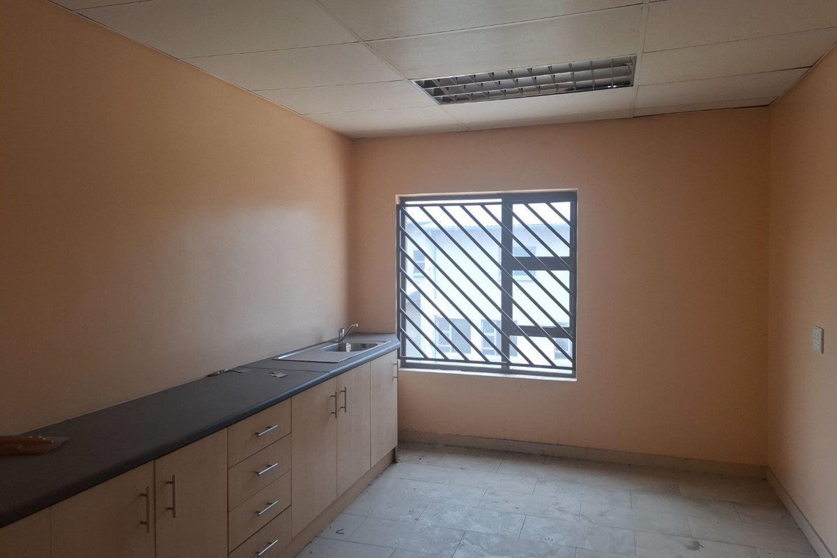 A functional kitchenette area featuring built-in cabinetry, a sink, and a window with security bars, suitable for office or utility use.