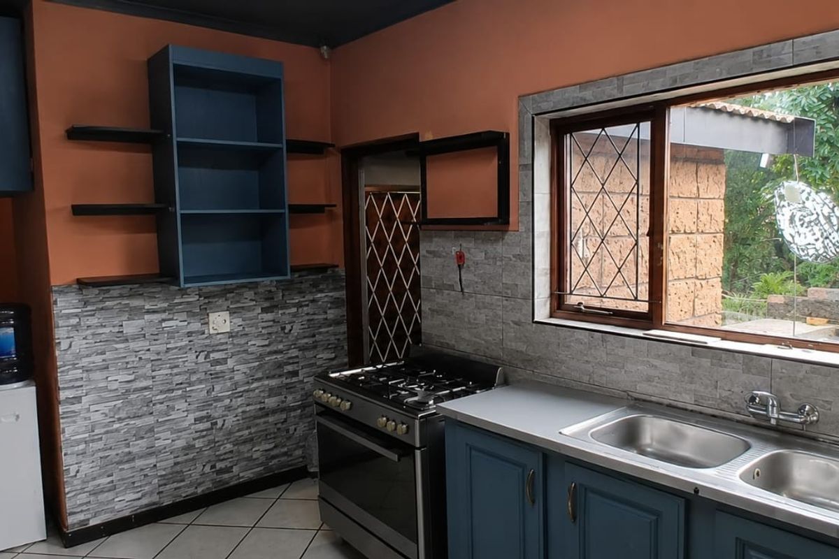 A functional kitchen featuring blue cabinetry, a stainless steel double sink, a gas stove, and stone-effect tiled splashbacks with a view to the garden.