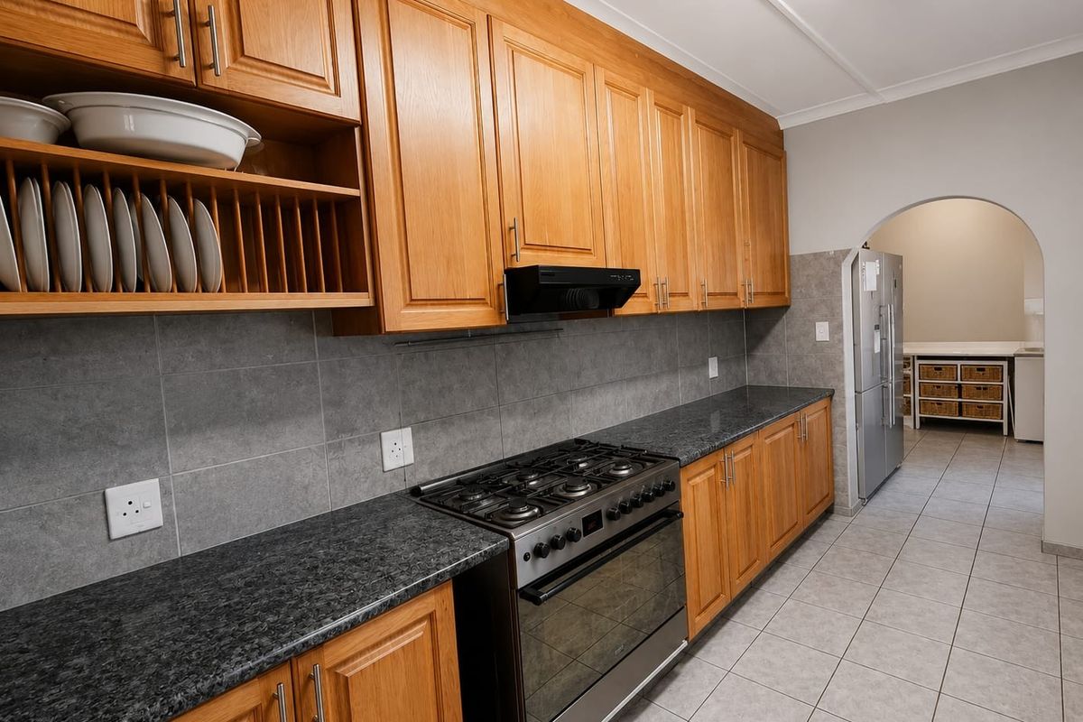 A functional kitchen featuring wooden cabinetry, granite countertops, a gas stove with extractor fan, and a tiled backsplash, leading into a secondary prep area.