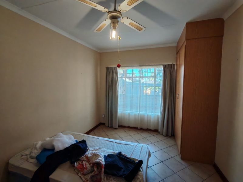 A bedroom featuring built-in wooden cupboards, tiled flooring, and a ceiling fan, offering a functional space with natural light.