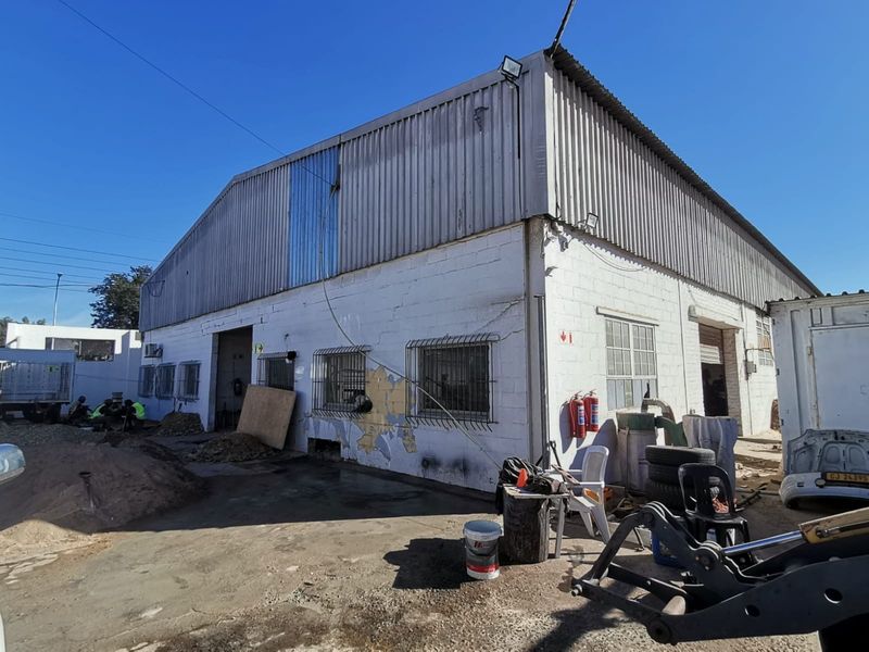 Exterior view of an industrial warehouse building featuring corrugated metal roofing, masonry walls, security bars on windows, and external fire extinguishers.
