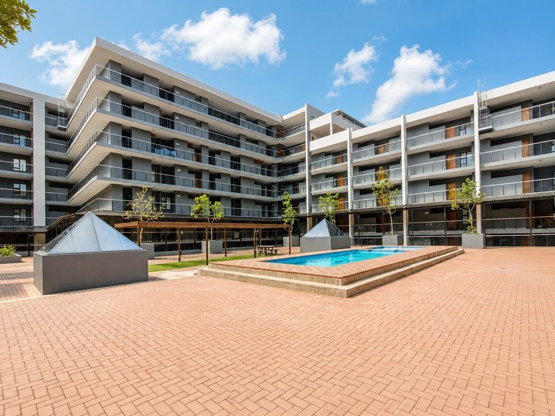A well-maintained apartment complex courtyard featuring a central swimming pool, paved terrace area, and modern multi-story building architecture with private balconies.