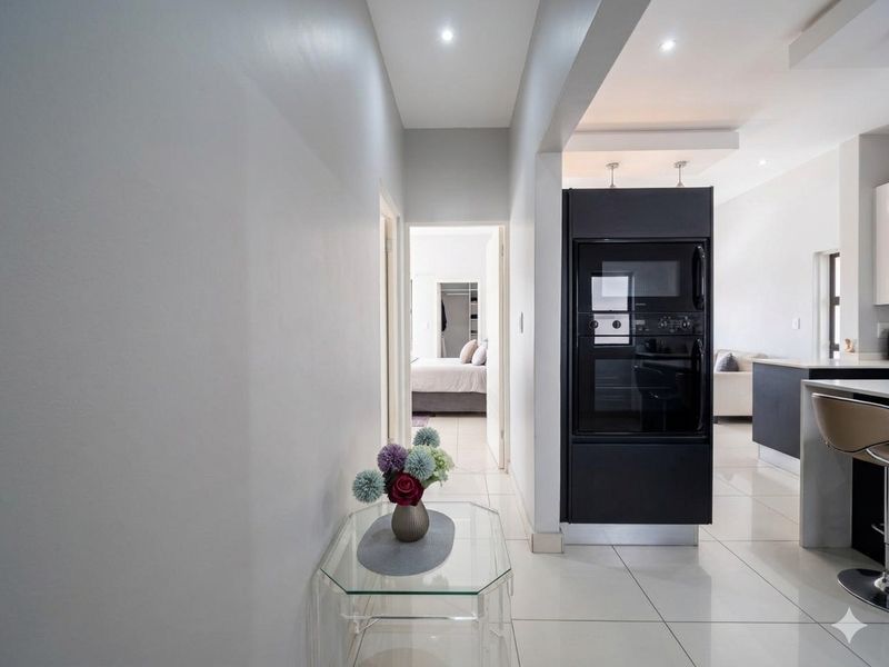 A bright hallway leading to a bedroom, featuring modern tiled flooring, recessed ceiling lighting, and a built-in black oven tower unit.