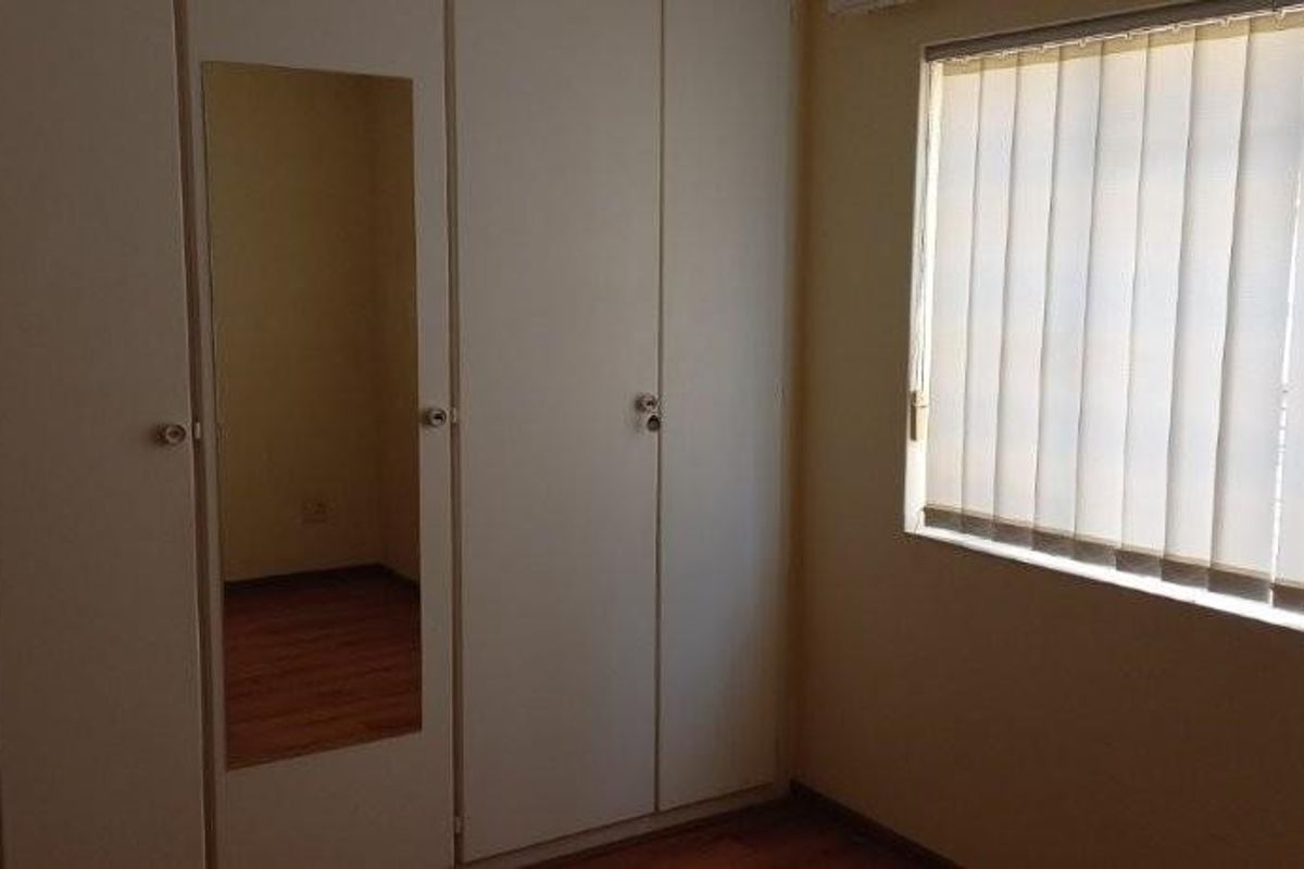 A bedroom featuring built-in cupboards with a mirrored panel and vertical blinds covering the window, finished with wooden flooring.
