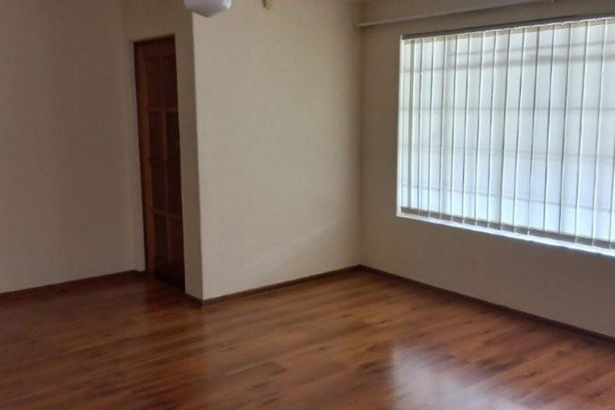 A spacious, empty living room featuring warm-toned laminate flooring, neutral walls, and large windows fitted with vertical blinds.