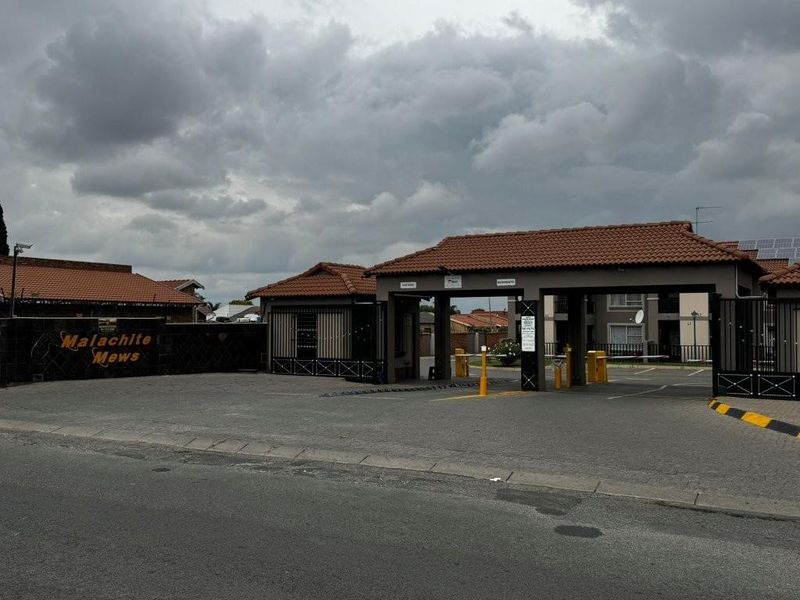The entrance to Malachite Mews apartment complex, featuring a gated security access point with tiled roof structures and paved driveway.