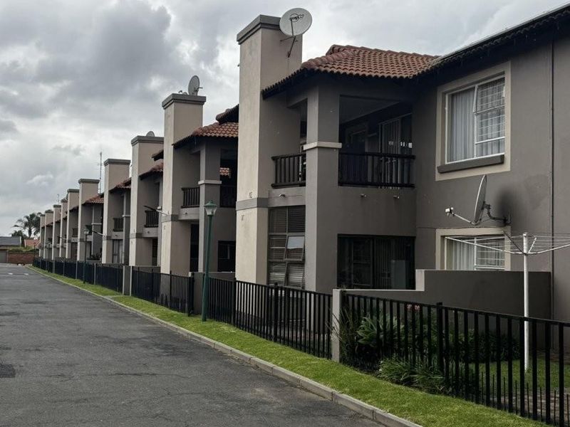 Exterior view of a multi-unit apartment complex featuring tiled roofs, private balconies, and ground-level fenced garden areas along a paved access road.
