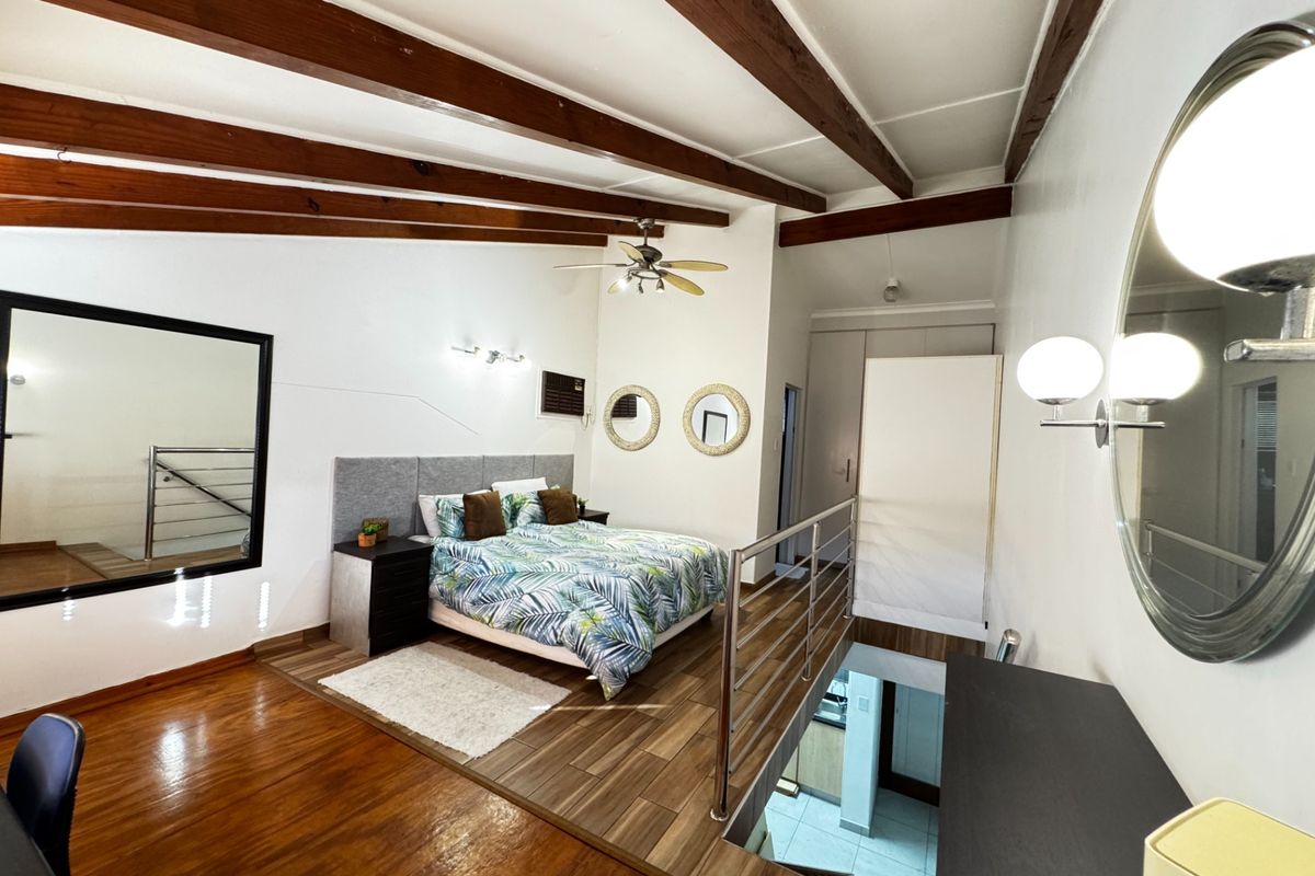 A mezzanine bedroom featuring exposed timber ceiling beams, wood-look flooring, and a stainless steel balustrade overlooking the lower level.