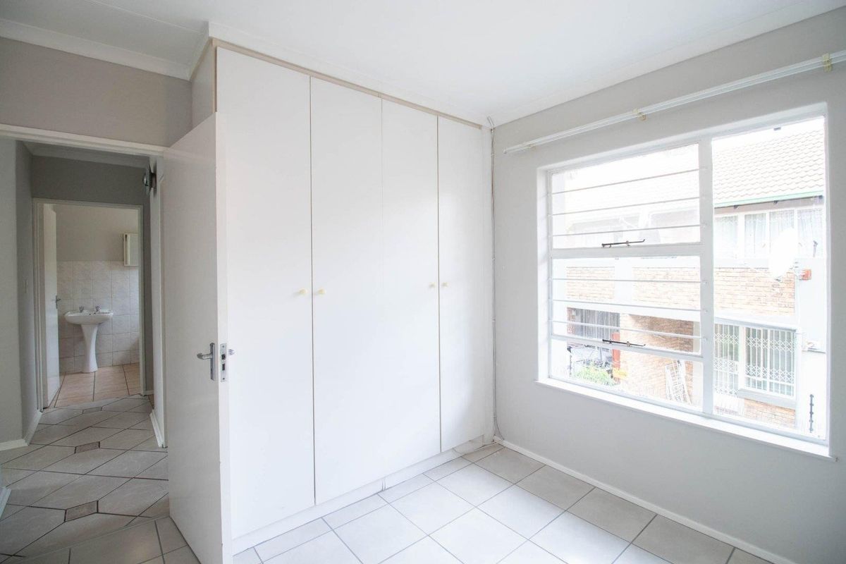 A bright bedroom featuring built-in cupboards, tiled flooring, and a window with security bars, with a view into the adjacent bathroom.