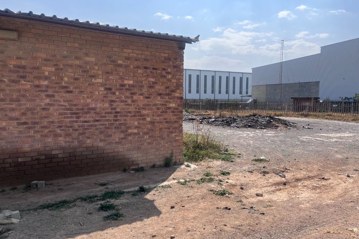 A view of a vacant residential plot featuring an existing brick structure and an unpaved, undeveloped yard area with visible debris.