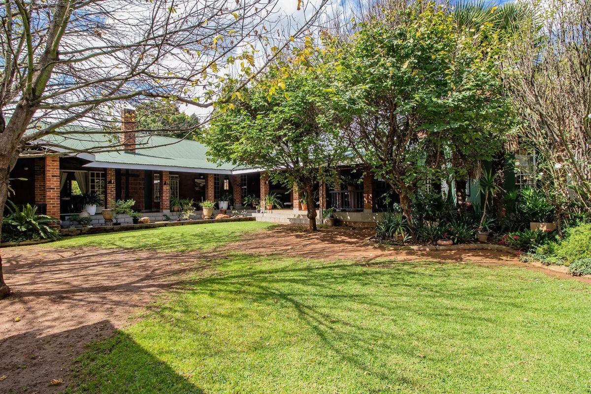 A spacious rear garden featuring a well-maintained lawn, mature trees, and a covered patio area with brick pillars attached to the main house.