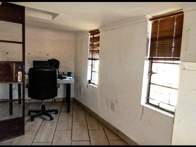A functional office space featuring tiled flooring, steel frame windows with timber blinds, and a built-in shelving unit, requiring cosmetic updates to walls.