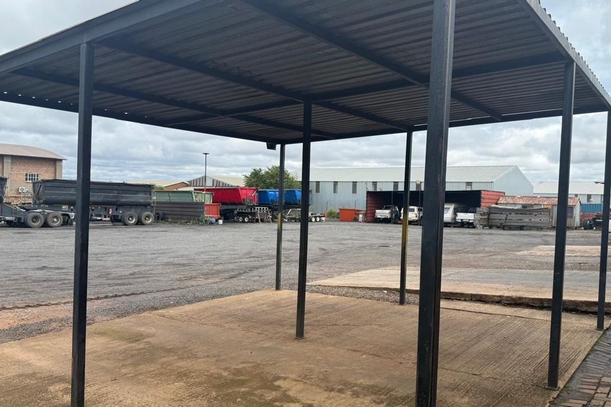 An open-sided industrial carport structure with a metal roof, providing covered outdoor space within a paved industrial yard.