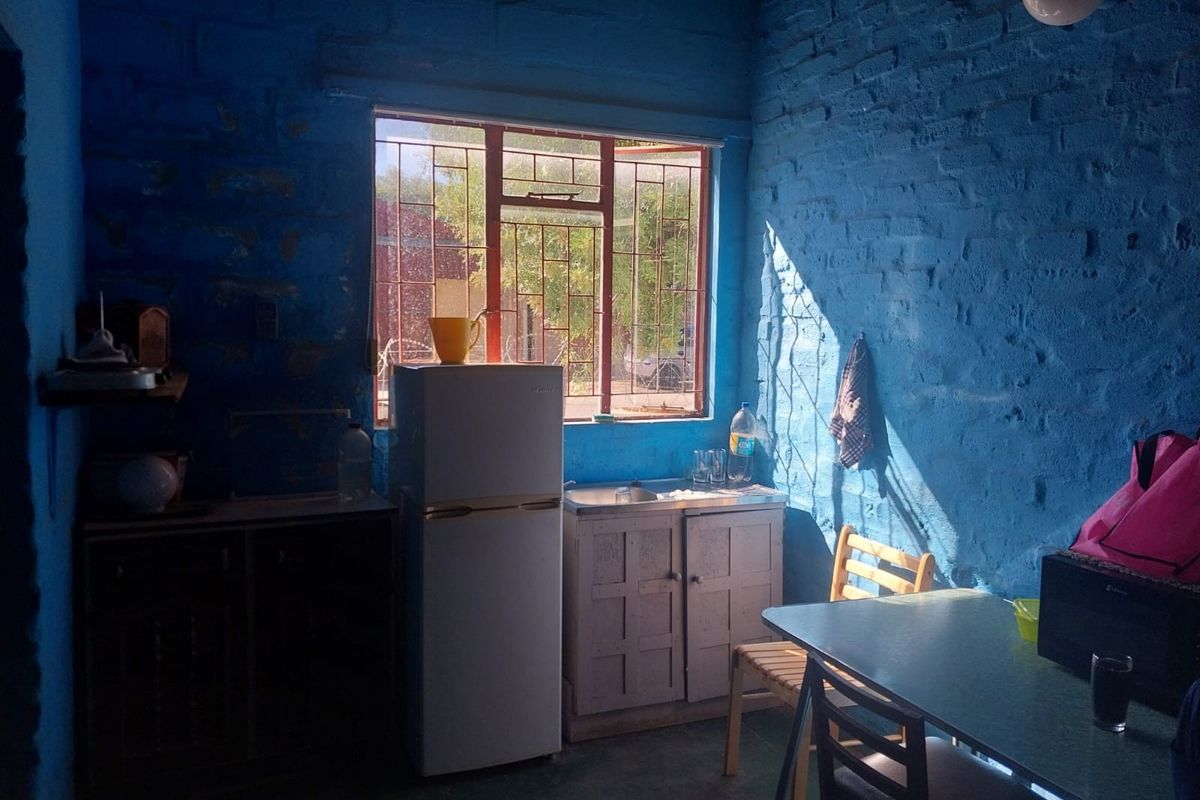 A basic kitchen area featuring exposed brick walls painted blue, a small sink unit, and a window with security bars.