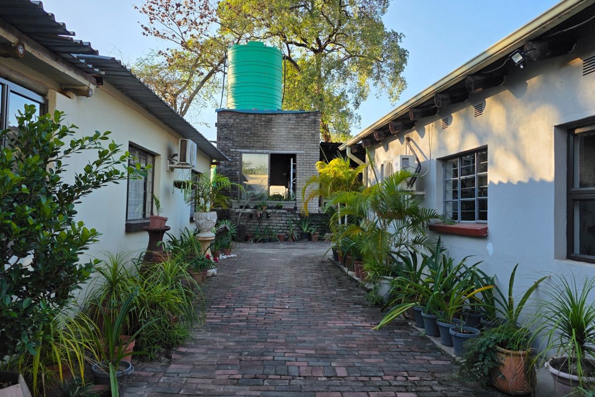 A paved courtyard area situated between two farm buildings, featuring a prominent elevated water tank and established garden borders.
