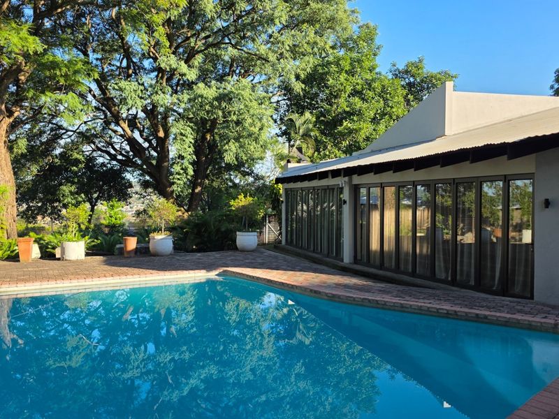 A well-maintained swimming pool area featuring brick paving, mature trees, and large glass sliding doors leading into the main residence.