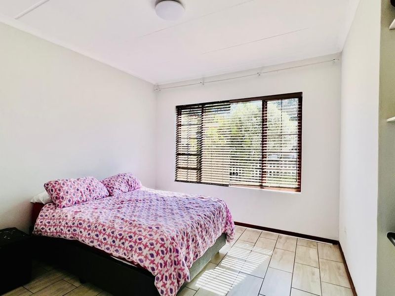 A bright bedroom featuring tiled flooring, built-in shelving, and a large window fitted with wooden venetian blinds.