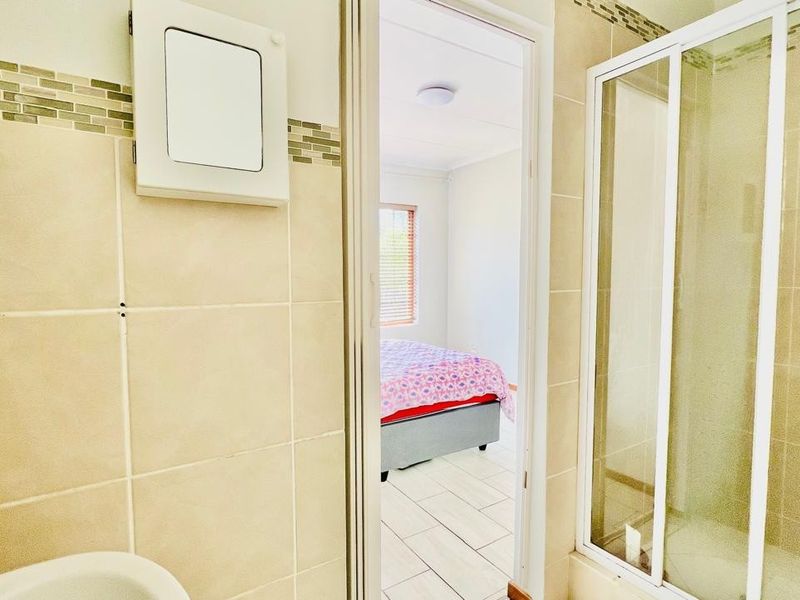 A view from a tiled bathroom into an adjacent bedroom, featuring a glass-enclosed shower and neutral wall tiling.