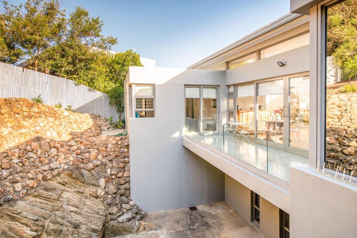 Modern rear exterior featuring a glass balustrade balcony, large sliding doors, and a natural stone retaining wall set against a landscaped hillside.