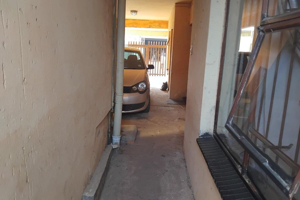 A narrow exterior walkway or side passage featuring concrete flooring, exterior wall finishes, and access to a parking area.