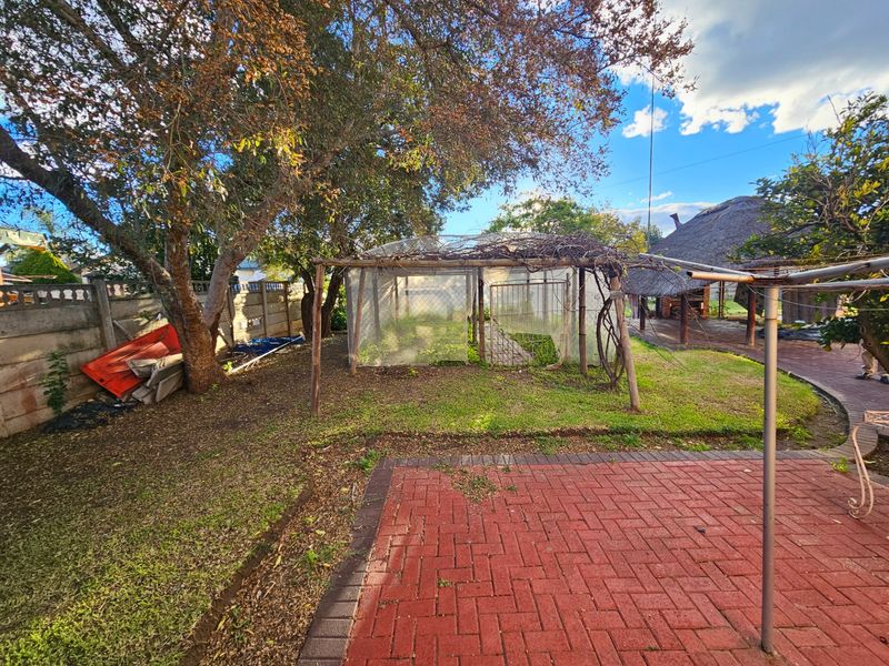 A spacious backyard featuring brick paving, a mature tree, and a garden trellis structure, with a thatched roof area visible in the background.