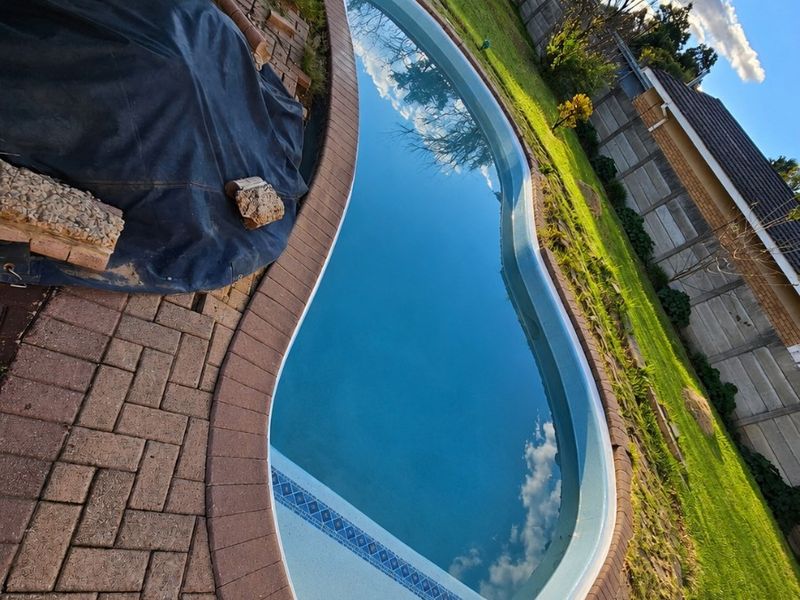 A kidney-shaped swimming pool set within a paved patio area, featuring a surrounding lawn and secure precast concrete boundary walling.