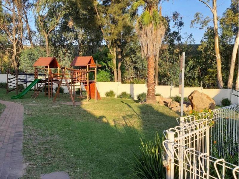 A communal garden area featuring a children's wooden playground structure, manicured lawn, mature palm tree, and secure perimeter walling with electric fencing.
