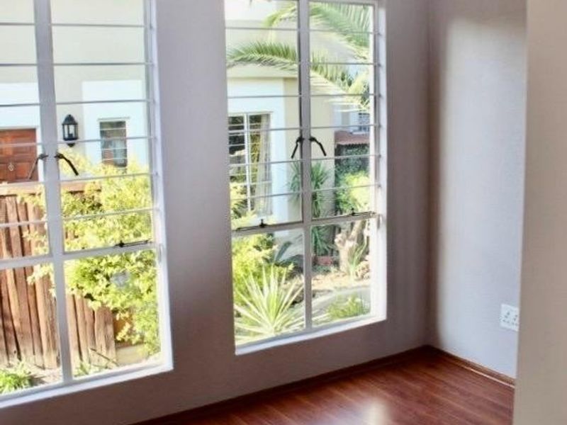 A bright, empty bedroom featuring warm-toned laminate flooring, neutral walls, and large windows with security bars overlooking the garden.