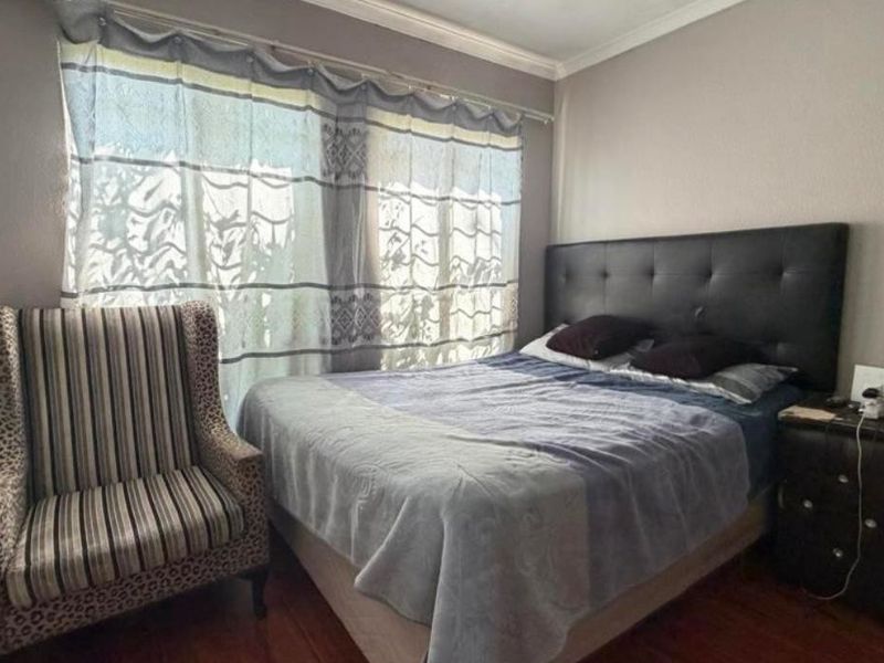 A bedroom featuring wooden flooring, neutral wall tones, and a large window with curtains, providing a comfortable and functional sleeping space.