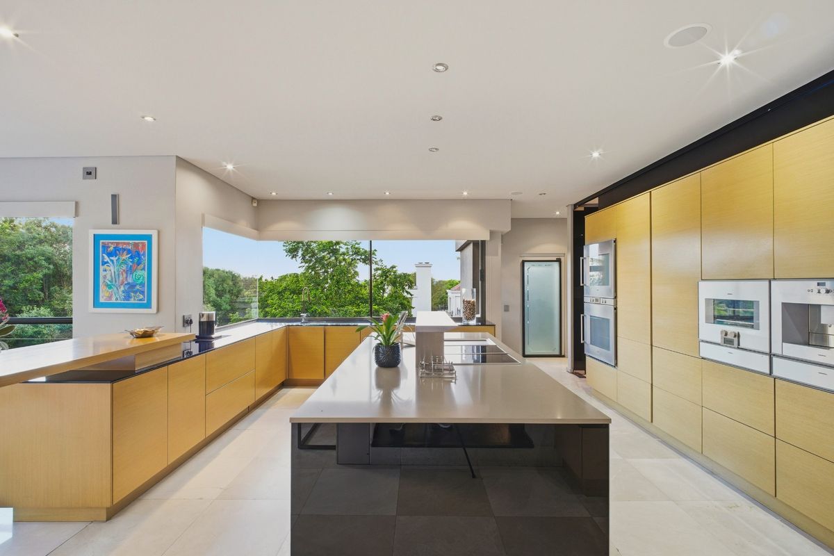 Modern kitchen featuring light wood cabinetry, a large central island with stone countertops, integrated appliances, and expansive windows overlooking greenery.